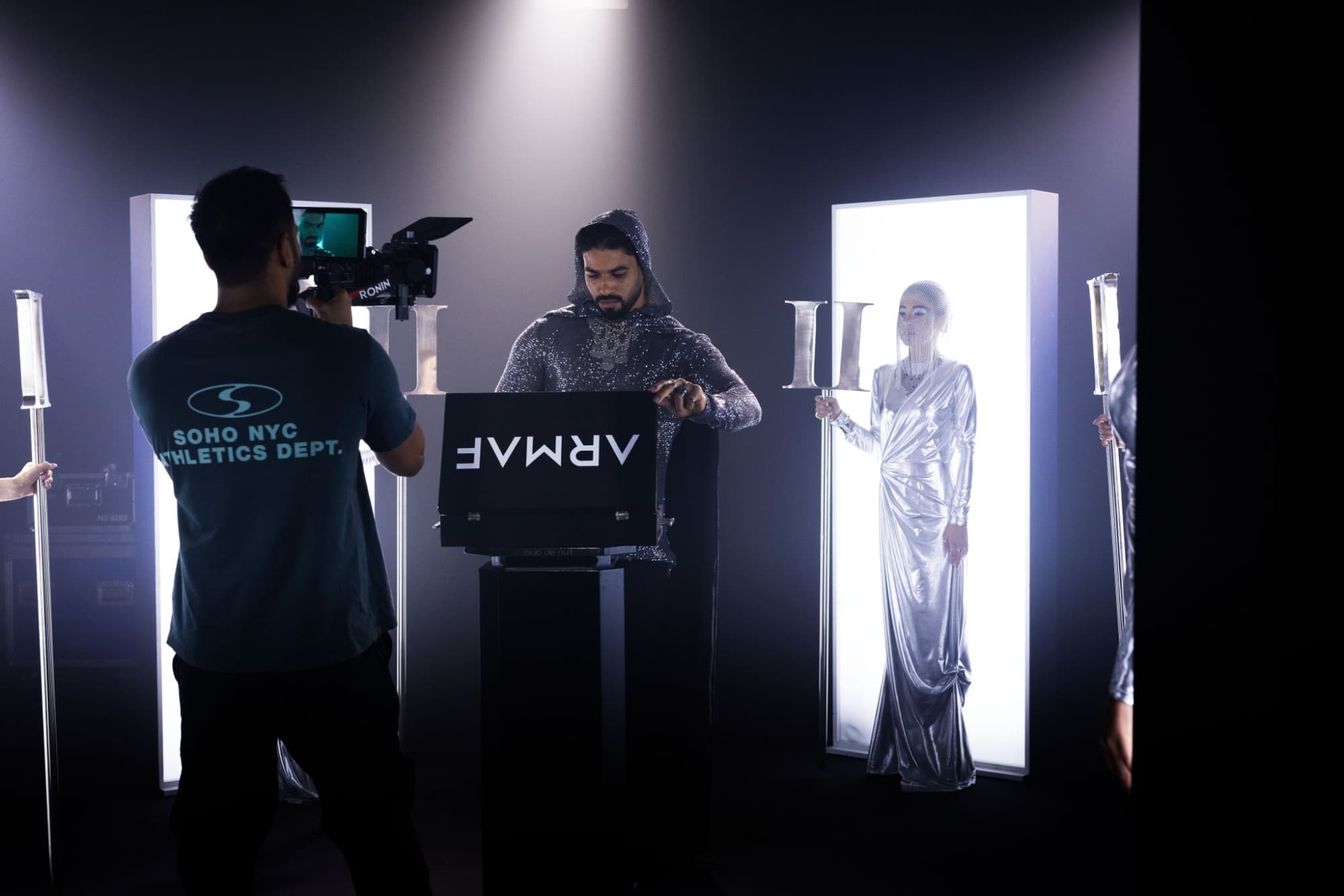 On the Armaf Precieux II set — a camera operator with a Ronin rig films the hooded lead at the ARMAF box, between light panels and a model in silver